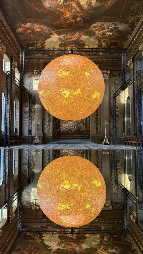 Luke Jerram’s Helios Exhibition at the Painted Hall in Greenwich, featuring a glowing Sun sculpture suspended beneath the stunning Baroque ceiling artwork.