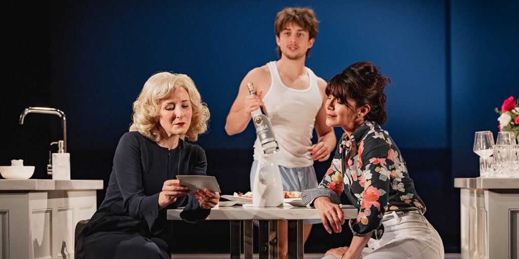 Dorothea Myer-Bennett, Gabriel Howell, and Caroline Catz in a tense kitchen scene from What We Talk About When We Talk About Anne Frank, with Howell holding a bottle of vodka.