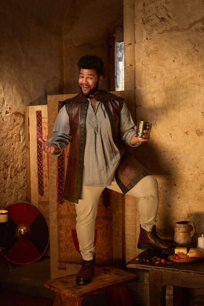 Emile Ruddock as Roland in A Knight’s Tale The Musical, smiling and holding a tankard in a rustic medieval setting.