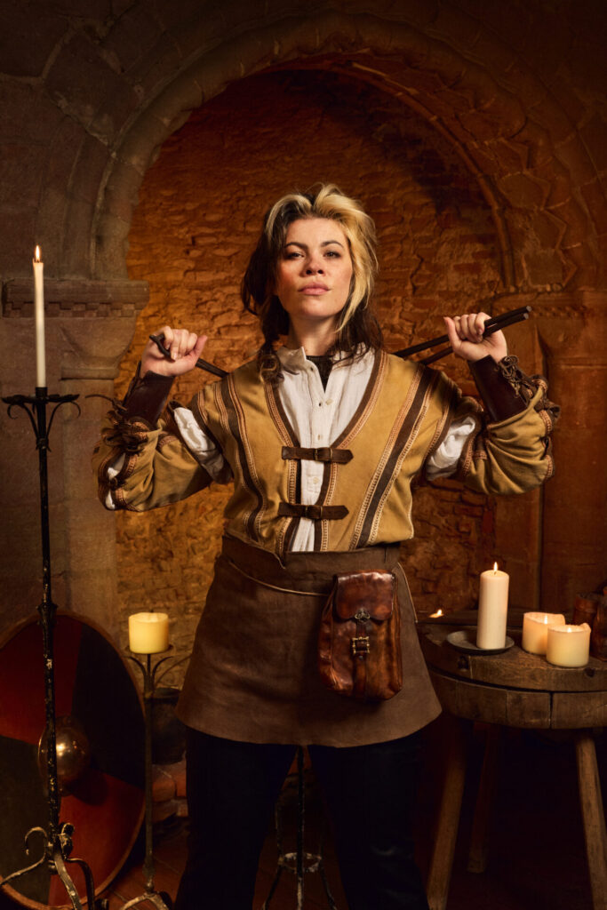Emily Benjamin as Kate in A Knight’s Tale The Musical, confidently holding tools with candles and medieval decor in the background.