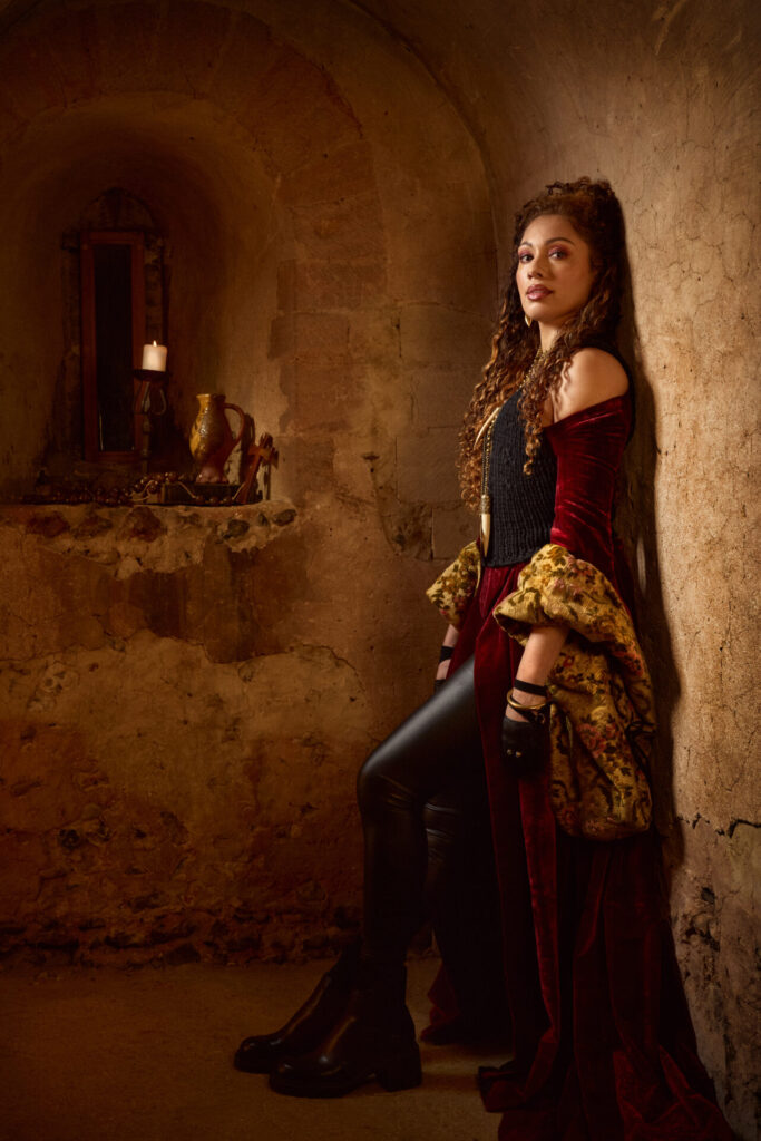 Meesha Turner as Jocelyn in A Knight’s Tale The Musical, leaning against a stone wall in a red velvet dress with gold detailing.